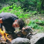 Man attempts to start a campfire surrounded by rocks in a lush green forest.