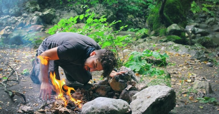 Man attempts to start a campfire surrounded by rocks in a lush green forest.