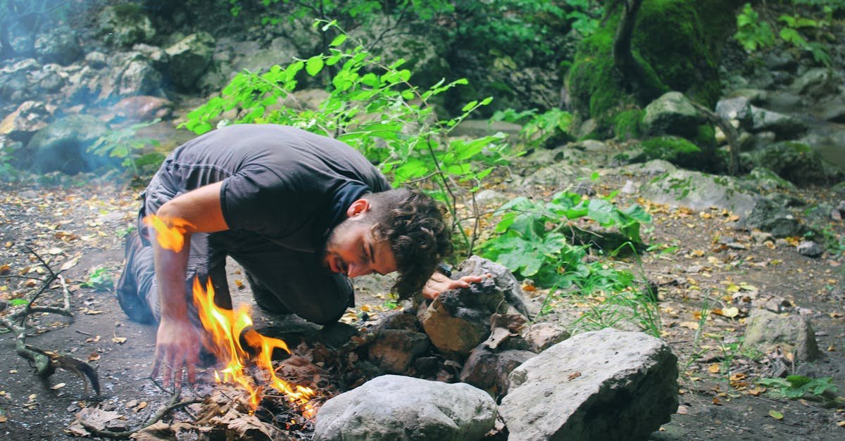 Man attempts to start a campfire surrounded by rocks in a lush green forest.