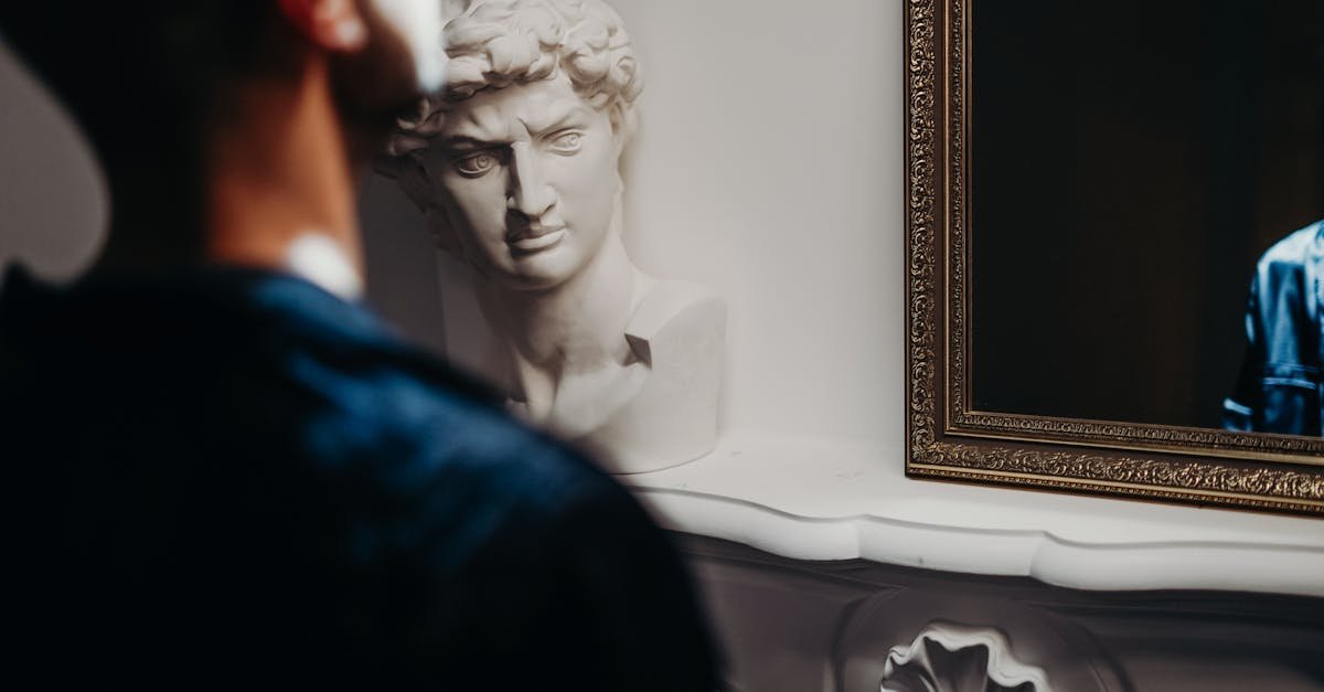 A man gazes into a mirror beside a classic sculpture in a sophisticated room.