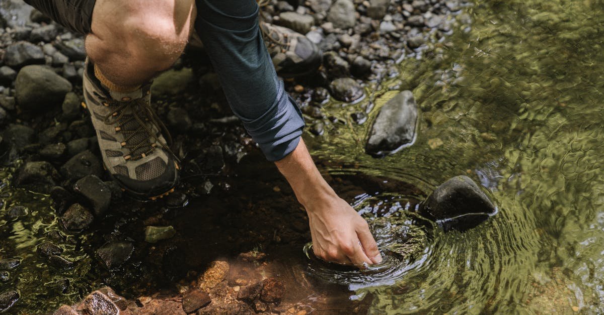 A man crouches down to touch a clear stream surrounded by rocks in a natural setting.